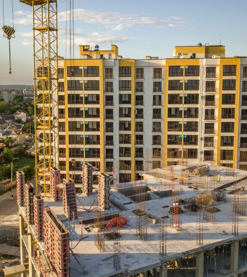 Apartment or office tall building under construction. Brick walls, glass windows, scaffolding and concrete support pillars. Tower crane on bright blue sky copy space background.